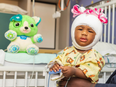 A toddler wearing a kid-friendly cranial head wrap sits in a crib near a stuff animal.